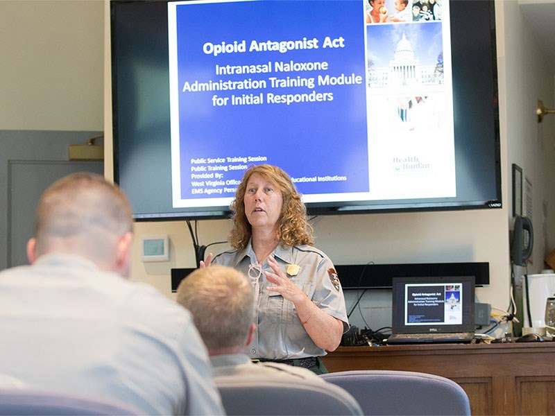 A person in a park ranger uniform stands towards the front of a room and in front of a screen with a presentation slide projected on it. The screen reads, “Opiod Antagonist Act.” There appears to be multiple other individuals dressed in park ranger uniforms in the audience, facing away from the camera.