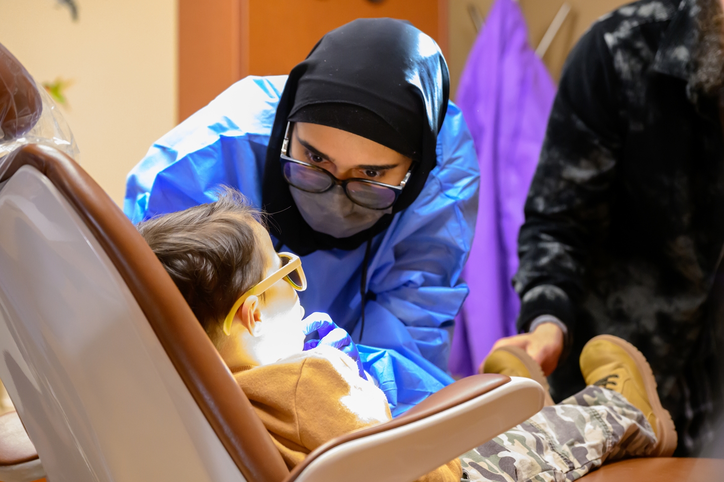 A young child wearing yellow protective glasses lies on a dental chair while a female dentist in a black hijab, blue gown, and mask examines their mouth.