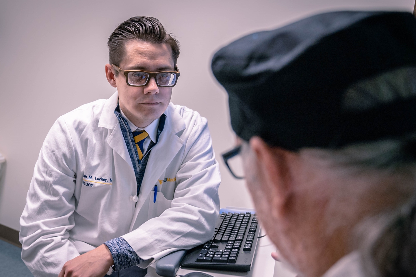 A young male doctor with glasses and a white lab coat sits at a desk facing an older patient, whose back is to the camera, with a computer keyboard and mouse visible on the desk.