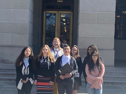 A group of seven students smile at the camera as they stand outside of a building on its steps.