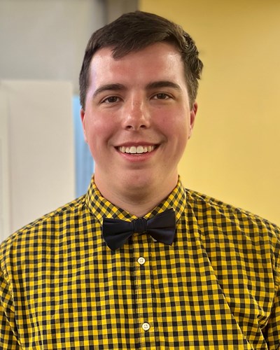 Ryan poses while wearing a gold and blue plaid button-down shirt and black bowtie. He is in a multipurpose room at the Health Sciences Center.