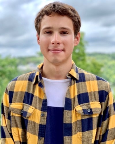 Alex smiles for the camera, wearing a gold and blue plaid button-down shirt. Behind him are some wooded hills and cloudy skies.