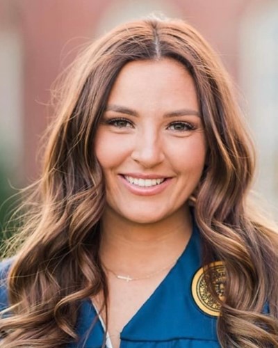Caylie stands smiling and looking at the camera in her blue graduation gown. She is standing in the middle of Woodburn Circle with Woodburn Hall behind her.