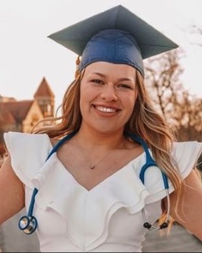 Danielle is seen smiling, wearing a white dress and blue graduation cap with a blue stethoscope draped around her neck. She is standing on the pedestrian bridge of the sunset-lit downtown campus of WVU.