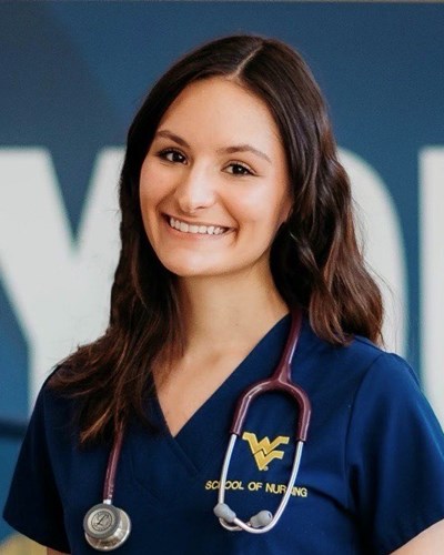 Stacy, wearing blue WVU School of Nursing scrubs and a stethoscope, smiles as she poses in front of the mural inside the Pylons lobby of the Health Sciences Center in Morgantown.