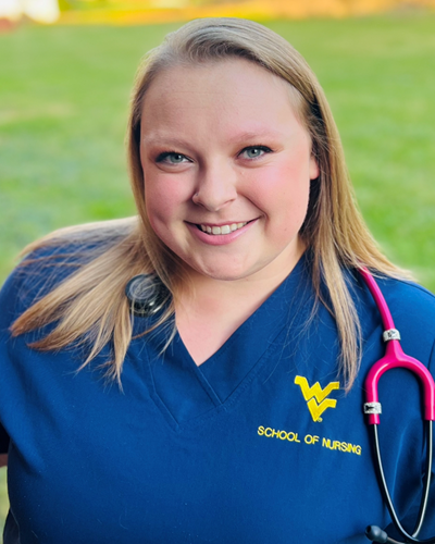 Rylee smiles while standing in a grassy area and wearing blue School of Nursing scrubs and a pink stethoscope draped over the back of her neck.