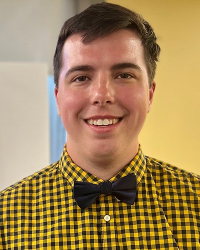 Ryan smiles for the camera. He is wearing a gold and dark gray plaid button-down shirt with dark gray bow tie.