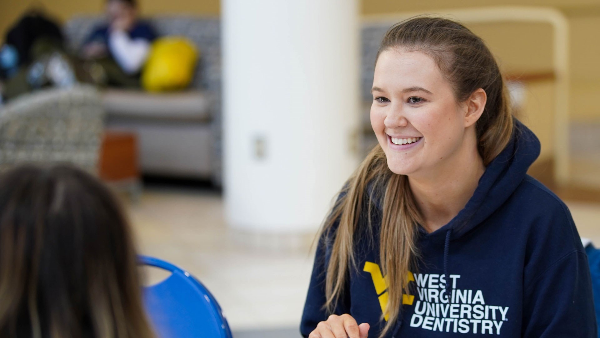 A person with long, brown hair smiles as they appear in the Pylons common area of the Health Sciences Center. They are wearing a dark blue West Virginia University hooded sweatshirt.