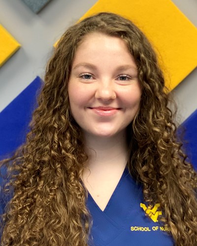 Hanna smiles for the camera, while wearing blue WVU School of Nursing scrubs and standing in front of a gray wall that has gold- and blue-colored and rotated square panels.