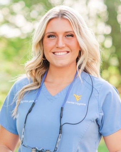 Leigh Ann smiles for the camera while standing outside amongst trees and wearing light blue WVU Dental Hygiene scrubs and corded dental spectacles around her neck.