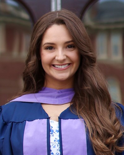 Kristen smiles while wearing a dark blue graduation gown lined with purple, and a purple sash around her neck. She is standing outside a doorway of a building on Woodburn Circle.
