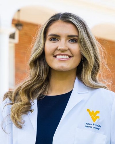 Lauren is seen smiling and wearing a white WVU Dental Hygiene coat over a navy blue dress. She is standing in front of a brick classroom building on the Downtown Campus.