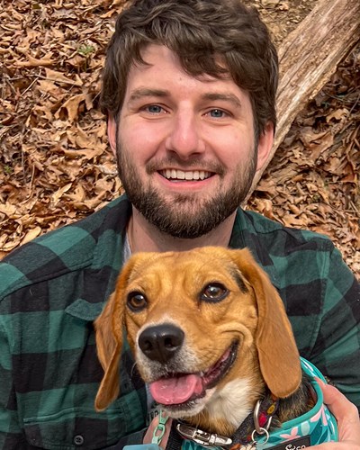 Ryan is seen smiling for the camera while sitting on a bench in a wooded area. He is wearing a dark green plaid flannel shirt, and has a dog sitting on his lap.