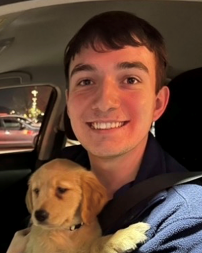 Coby smiles for the camera, while sitting in the front seat of a vehicle while holding a golden retriever puppy.