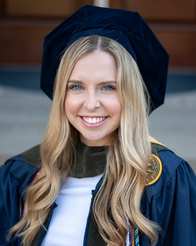 Megan is seen smiling, sitting on the steps in front of a WVU building, wearing a cap and gown.