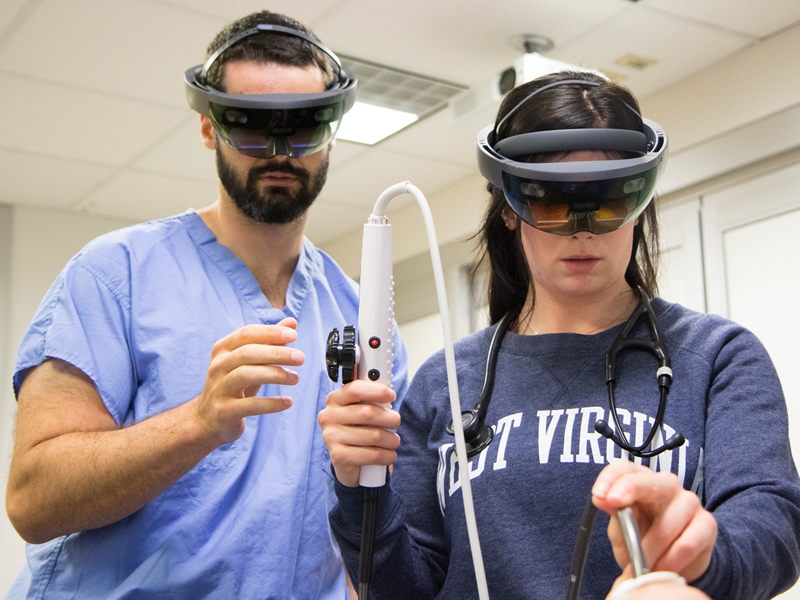 Two individuals wearing augmented reality training headsets engage in an ultrasound medical training simulation. One holds a medical device while the other, dressed in scrubs, observes and assists.