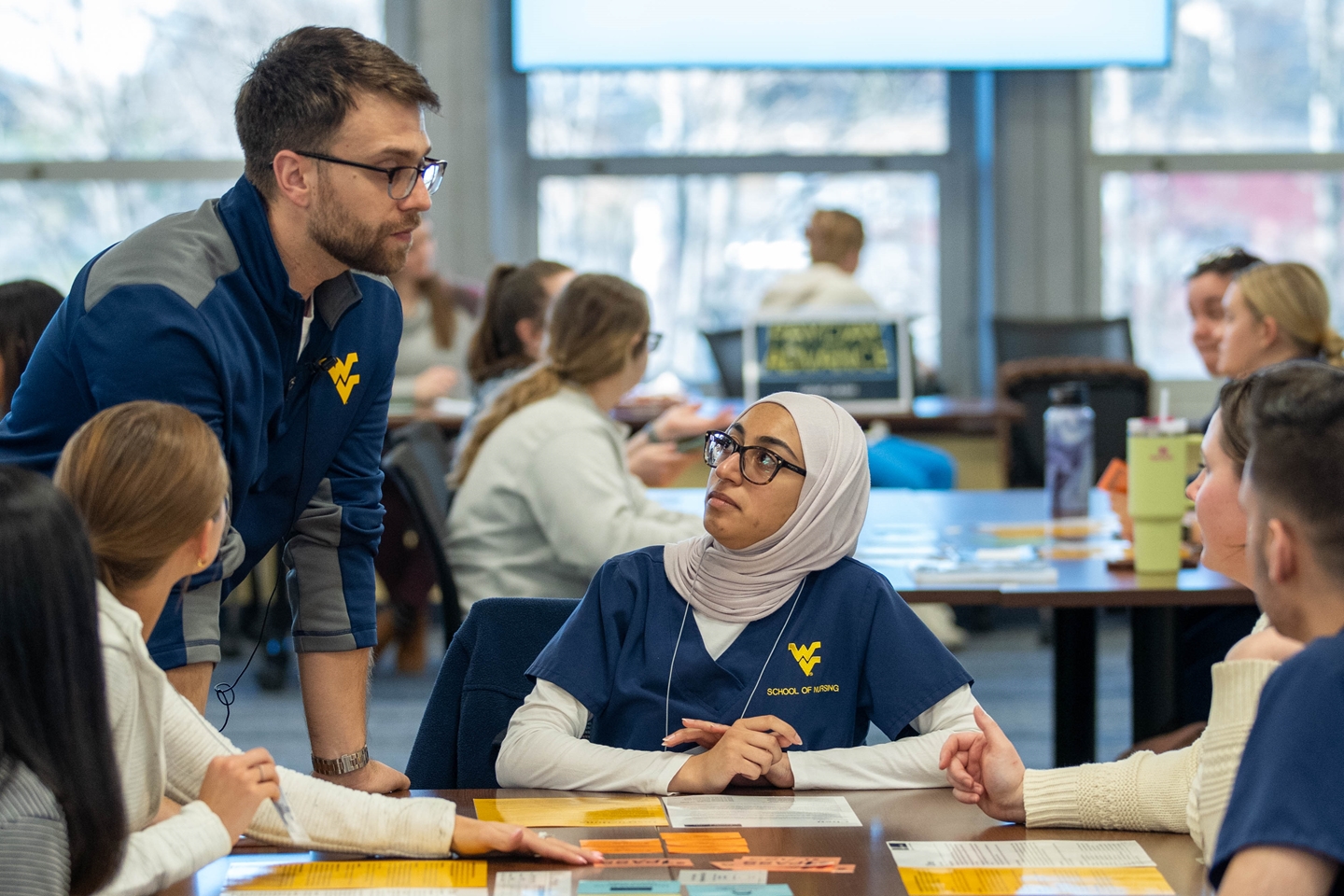 A WVU instructor in a blue jacket speaks with students. A woman in a hijab and navy scrubs looks up as papers cover the table in a bright room.