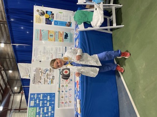 A person dressed in a white coat and scrubs by a display table and poster at the Mon county fair.