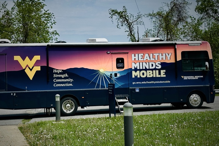 A large, dark blue bus parked in a lot with trees in the background. The side of the bus features a vibrant wrap of a mountain sunset and the yellow West Virginia University logo. Large text on the side reads "HEALTHY MINDS MOBILE" alongside the words "Hope. Strength. Community. Connection."